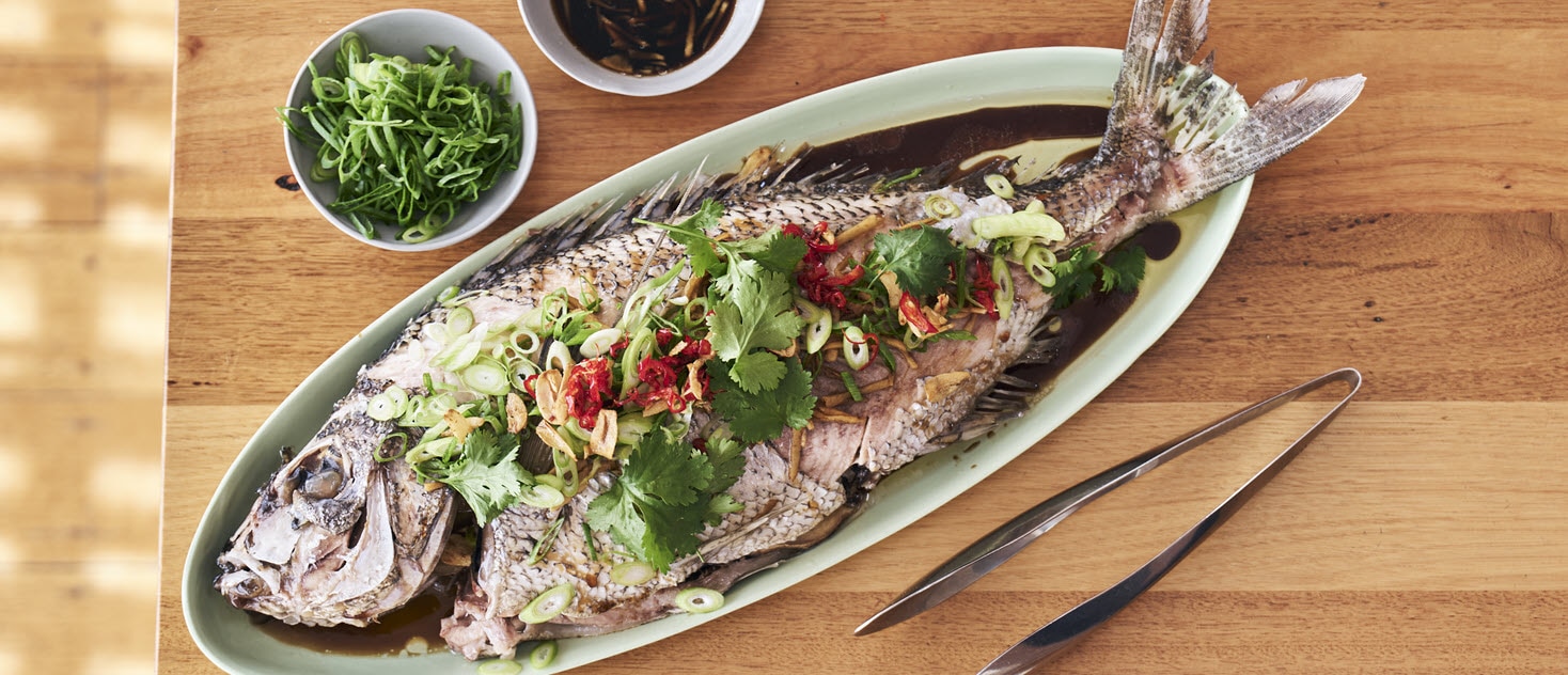 Steamed whole fish garnished with coriander, spring onions, and crispy garlic, served with a side of soy-chilli sauce and steamed rice.